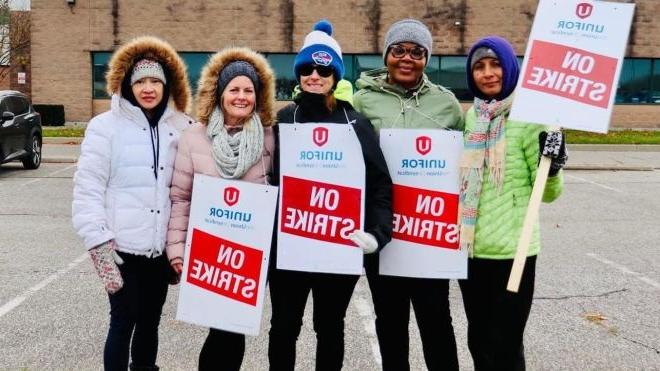Five women standing together outside holding red and white placards that read on strike with the uedbet新版官网 logo displayed on them in front of a the Magna building in Windsor, Ontario.
